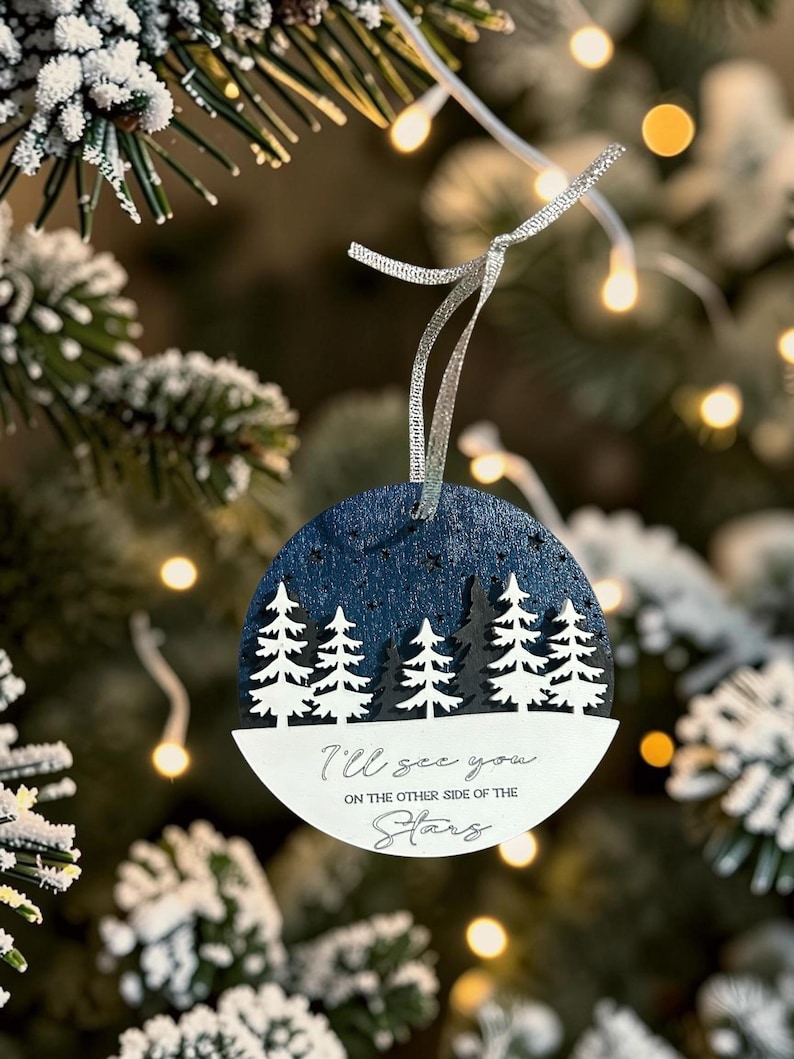 May include: A circular wooden ornament featuring a dark blue night sky with a forest of white pine trees. The lower white section displays the black text: I'll see you ON THE OTHER SIDE OF THE Stars. A silver ribbon is attached to the top for hanging.