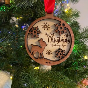Op de afbeelding: Houten kerstbal met een hert-silhouet en sneeuwvlok-uitsparingen. De bal heeft de tekst "My First Christmas Mackenzie Grace 2022" en hangt aan een rood lint. Het hangt in een kerstboom.