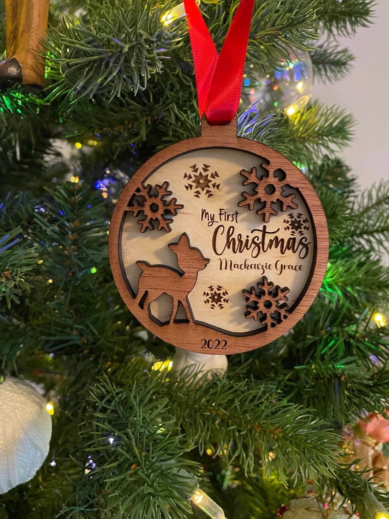 Op de afbeelding: Houten kerstbal met een hert-silhouet en sneeuwvlok-uitsparingen. De bal heeft de tekst "My First Christmas Mackenzie Grace 2022" en hangt aan een rood lint. Het hangt in een kerstboom.