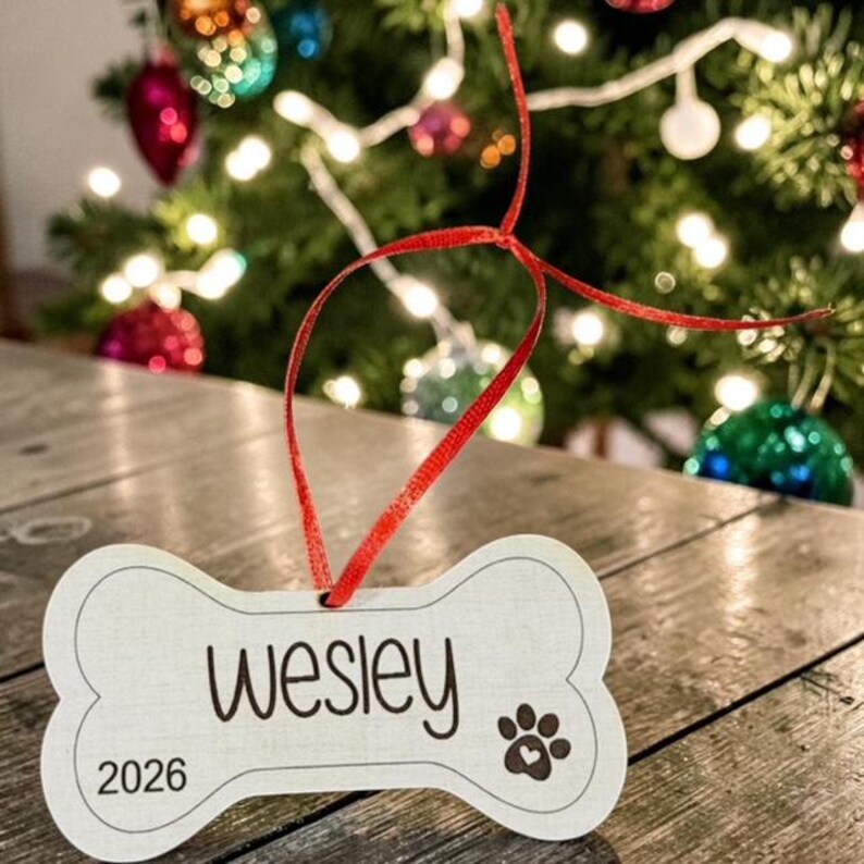May include: A white bone-shaped ornament featuring the name Wesley in black script, the year 2026, and a paw print icon with a small heart in the center. The ornament is suspended by a thin red ribbon tied in a loop at the top center.