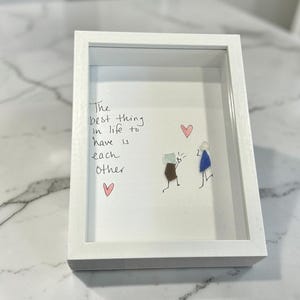 May include: A white shadow box frame containing a piece of sea glass art. The artwork features two figures made of sea glass, one in brown and one in blue, with hand-drawn details and two pink hearts. The text reads: The best thing in life to have is each other.