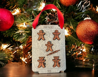 May include: A personalized Christmas ornament with the text "OUR FAMILY" at the top. The ornament features gingerbread people and dog shapes with names, and the year "2023" at the bottom. A red ribbon is attached for hanging.