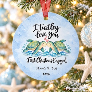 May include: A round ceramic Christmas ornament with a light blue watercolour background. It features two green sea turtles facing each other with three small red hearts above them. Text reads: I turtley love you, First Christmas Engaged, Jenna & Joe, 2026.