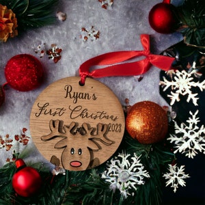 May include: A wooden Christmas ornament featuring the text "Ryan's First Christmas 2023" and a reindeer design. The round ornament has a red ribbon. The image includes red and gold baubles, snowflakes, and festive greenery.