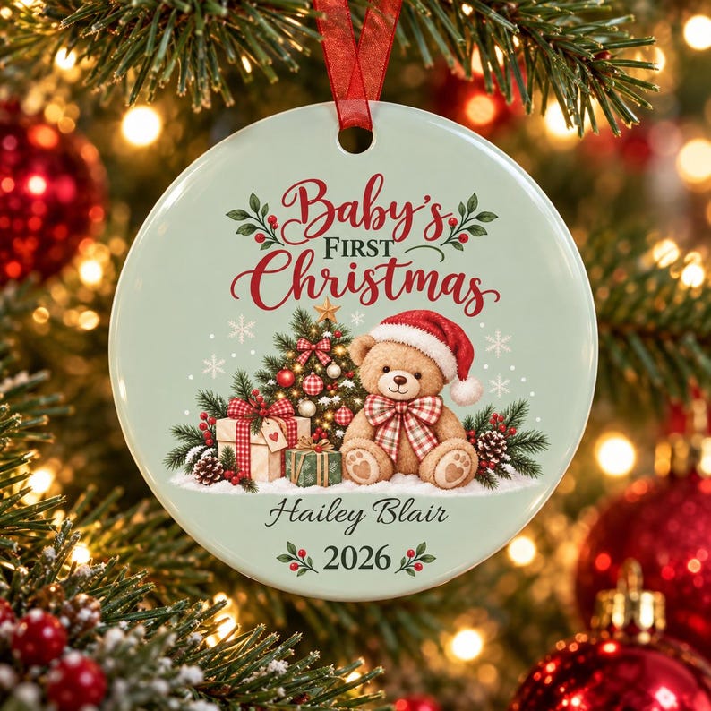 May include: A round, pale green ceramic Christmas ornament featuring a teddy bear wearing a red Santa hat, a decorated Christmas tree, and wrapped gifts. The text reads Baby's First Christmas, Hailey Blair, and 2026 in red and black script.