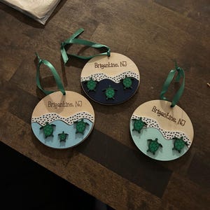 May include: Three wooden ornaments with green ribbon hangers. Each ornament features a light wood base with a painted ocean scene and green sea turtles. The text "Brigantine, NJ" is printed on each ornament.