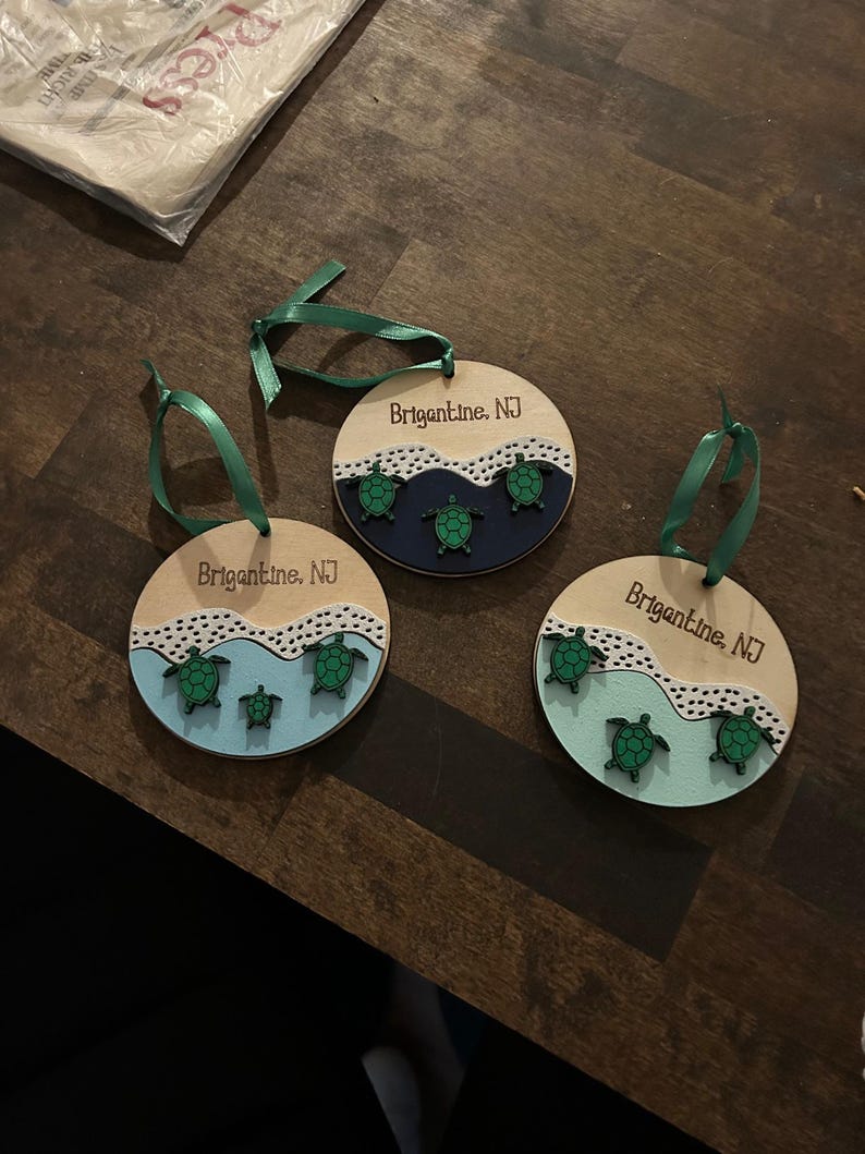 May include: Three wooden ornaments with green ribbon hangers. Each ornament features a light wood base with a painted ocean scene and green sea turtles. The text "Brigantine, NJ" is printed on each ornament.