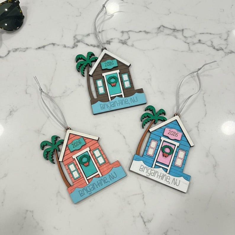 Puede incluir: Tres adornos de madera con forma de casa de playa y cordones plateados. Cada uno incluye una palmera, una corona en la puerta y el texto 2026 y Brigantine, NJ. Las casas son de color marr&oacute;n, coral y azul con bordes blancos y bases celestes.