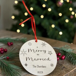 May include: A round, light-colored wooden ornament featuring a red ribbon hanger. The surface is engraved with the text Merry + Married, The Sheridans, and 09.12.26, surrounded by various snowflake designs in a dark, etched font style.