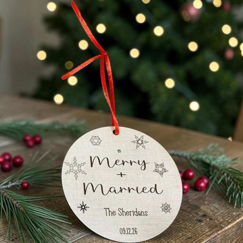 May include: A round, light-colored wooden ornament featuring a red ribbon hanger. The surface is engraved with the text Merry + Married, The Sheridans, and 09.12.26, surrounded by various snowflake designs in a dark, etched font style.