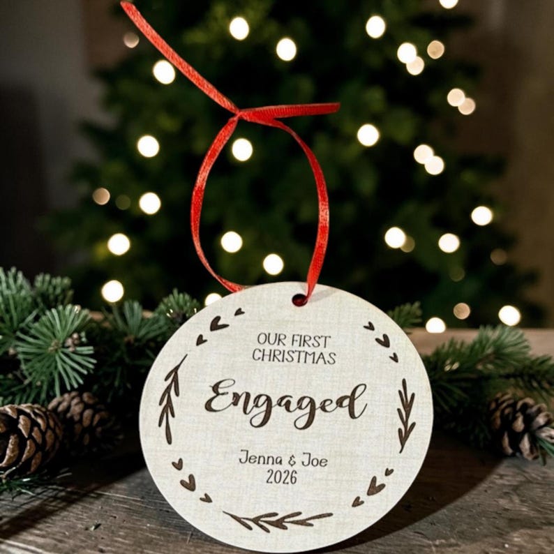 May include: A round, light-coloured wooden ornament featuring a red ribbon loop at the top. The text reads "OUR FIRST CHRISTMAS Engaged" with the names "Jenna & Joe" and the year "2026" engraved, surrounded by a border of small hearts and leaves.