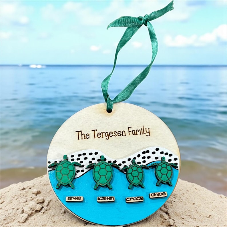 May include: A wooden family ornament with the text "The Tergesen Family". The ornament features four green turtles above names on a blue background. A green ribbon is attached for hanging. The ornament is on a sandy beach.