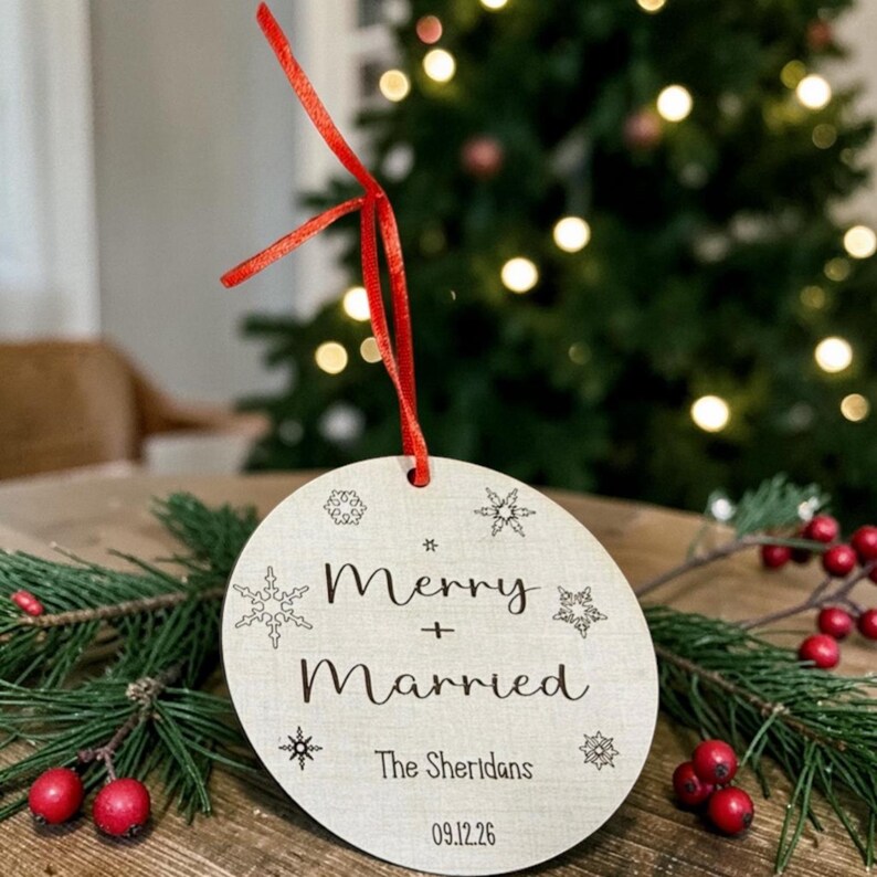 May include: A round, light-colored wooden ornament featuring a red ribbon loop at the top. The text "Merry + Married", "The Sheridans", and "09.12.26" is printed in black, surrounded by small, engraved snowflake designs of varying shapes.