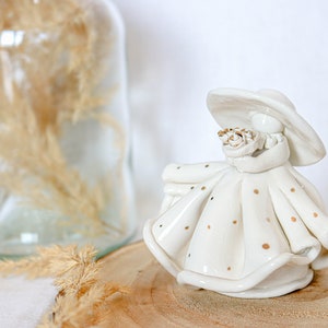 Może przedstawiać: Biała ceramiczna figurka kobiety w białej sukience w złote kropki i szerokim kapeluszu. Trzyma bukiet kwiatów w dłoniach.