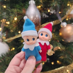 May include: Two small Christmas elf ornaments. One elf is wearing a blue outfit and hat with white trim, the other is wearing a red outfit and hat with white trim. The ornaments have large blue eyes and are set against a Christmas tree background.