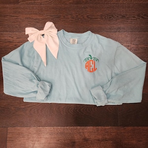 May include: A light blue long-sleeve shirt with a pumpkin and monogram design. The pumpkin is orange with green vines, and the monogram is also orange. A large white bow is placed on the left side of the shirt.