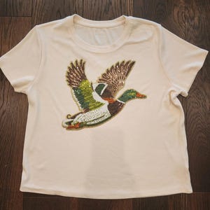 May include: Cream-colored short-sleeved t-shirt featuring a detailed sequined design of a flying duck. The duck has brown, green, and white feathers with gold accents. The shirt is made of a soft, ribbed fabric.