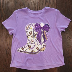 May include: A lilac-colored t-shirt featuring a graphic of a white cowboy boot adorned with footballs and stars. A purple bow with the word "TOUCHDOWN" is on the right side of the boot. The shirt is made of a ribbed fabric.