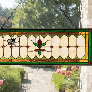 May include: A rectangular stained glass window panel with a bee and floral design. The panel features a green, gold, and red color scheme, with a chain for hanging. The window is set against a backdrop of a window and a garden.