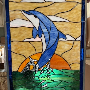 May include: Stained glass panel featuring a leaping dolphin in shades of blue and white against a sunset backdrop. The artwork includes green water and white clouds, framed by a blue border. A decorative piece.