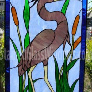 May include: A stained glass window depicting a blue heron standing in a marsh with cattails. The heron is brown with a long neck and beak. The background is a light blue sky.