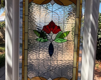 May include: A stained glass window with a red flower design. The flower is surrounded by green leaves and a blue oval. The window is framed in white.