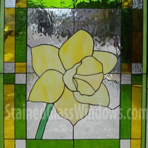 May include: A stained glass window featuring a yellow daffodil with green leaves against a background of green and yellow glass squares. The window is framed in a dark wood frame.