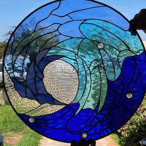 May include: A stained glass window depicting a blue and turquoise wave crashing against a white background. The wave is made up of many small pieces of glass, creating a textured effect.