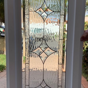 May include: A white framed stained glass window with a diamond pattern and textured glass. The window is set against a blurred background of greenery.