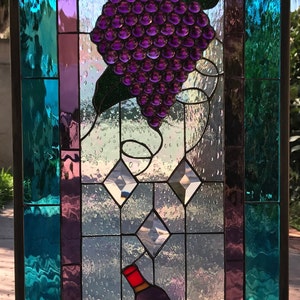 May include: A stained glass window depicting a grapevine with a bunch of purple grapes and a wine bottle. The background is a blue and purple iridescent glass.