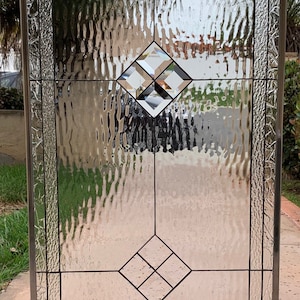 May include: Stained glass window with a textured, rippled surface. The design features geometric shapes, including diamonds and squares, with accents of yellow, green, and clear glass. The window is framed in a silver metal.