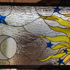 May include: Stained glass panel featuring a celestial design. The artwork depicts a yellow sun with a smiling face, blue stars, and a crescent moon against a textured, silver background. The panel is framed in black.