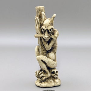 Peut inclure: Une petite figurine en céramique blanche d'une gargouille tenant un crâne. La gargouille a une expression menaçante et tient un crâne dans sa main droite.