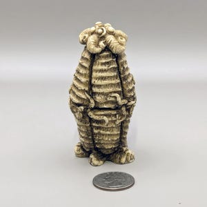 May include: A small, cream-colored ceramic figurine of a creature with a textured, segmented body and a crown of small, rounded shapes on its head.
