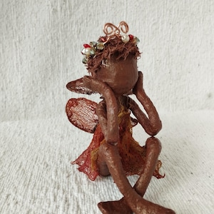 May include: A brown textured figurine of a seated creature with wings, wearing a copper wire crown adorned with small red, white, and green beads. The figure has a textured brown body, a brown head, and a skirt made of reddish-brown mesh fabric.