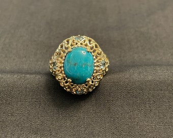 May include: A gold ring featuring a large turquoise stone set within a filigree design. The ring is embellished with smaller clear stones.