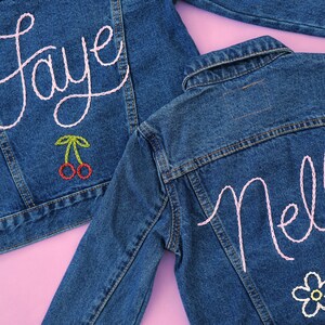 Puede incluir: Dos chaquetas vaqueras con nombres bordados en rosa, "Faye" y "Nell", en la espalda. La chaqueta con "Faye" tiene un par de cerezas rojas bordadas debajo del nombre. La chaqueta con "Nell" tiene una flor blanca bordada debajo del nombre.