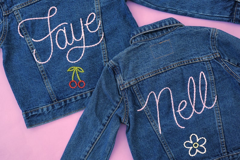 Puede incluir: Dos chaquetas vaqueras con nombres bordados en rosa, "Faye" y "Nell", en la espalda. La chaqueta con "Faye" tiene un par de cerezas rojas bordadas debajo del nombre. La chaqueta con "Nell" tiene una flor blanca bordada debajo del nombre.