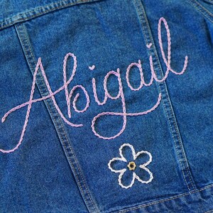 May include: A blue denim jacket with the name "Abigail" embroidered in pink thread on the back. A white flower with a brown center is embroidered below the name.