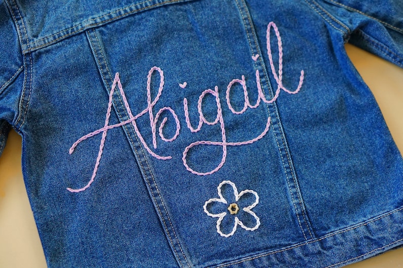 May include: A blue denim jacket with the name "Abigail" embroidered in pink thread on the back. A white flower with a brown center is embroidered below the name.