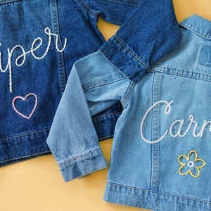 May include: Two denim jackets with embroidered names, one with the name "Piper" and a pink heart, the other with the name "Carmen" and a yellow flower.