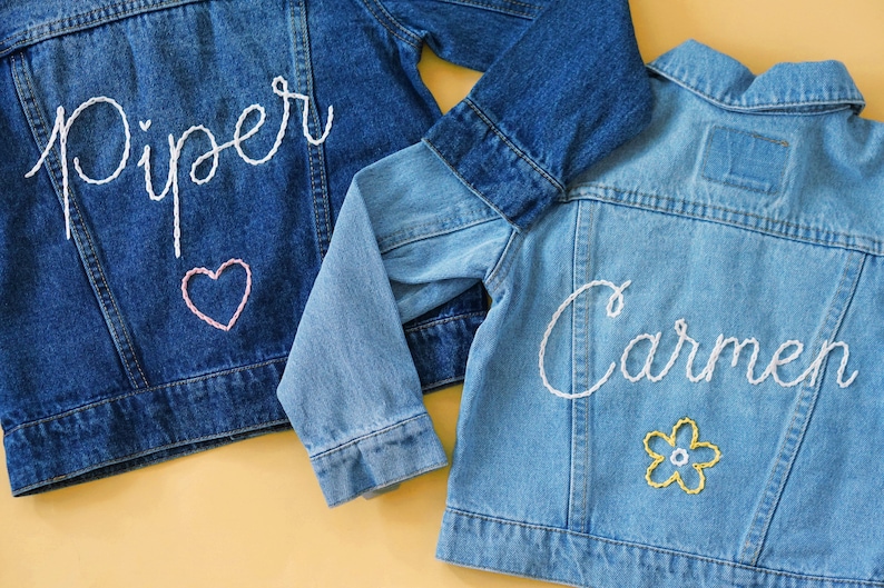 May include: Two denim jackets with embroidered names, one with the name "Piper" and a pink heart, the other with the name "Carmen" and a yellow flower.