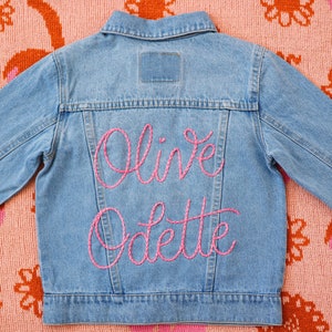 May include: A blue denim jacket with pink embroidery that reads "Olive Odette" on the back.