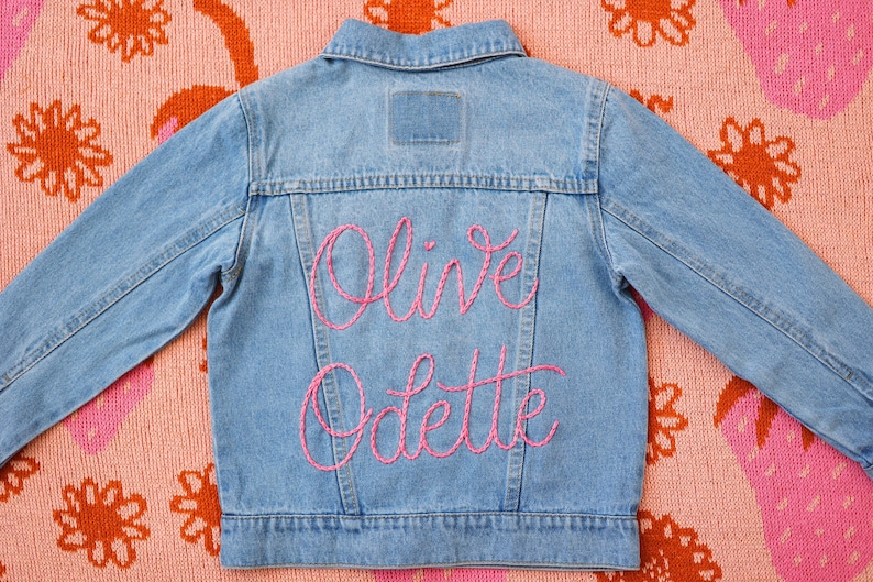 May include: A blue denim jacket with pink embroidery that reads "Olive Odette" on the back.