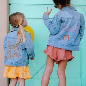 Puede incluir: Dos ni&ntilde;as peque&ntilde;as con chaquetas vaqueras con dise&ntilde;os bordados. La ni&ntilde;a de la izquierda lleva una falda amarilla con limones y el texto "Candy June" bordado en su chaqueta. La ni&ntilde;a de la derecha lleva una falda rosa y el texto "Drew" y un arco&iacute;ris bordado en su chaqueta.