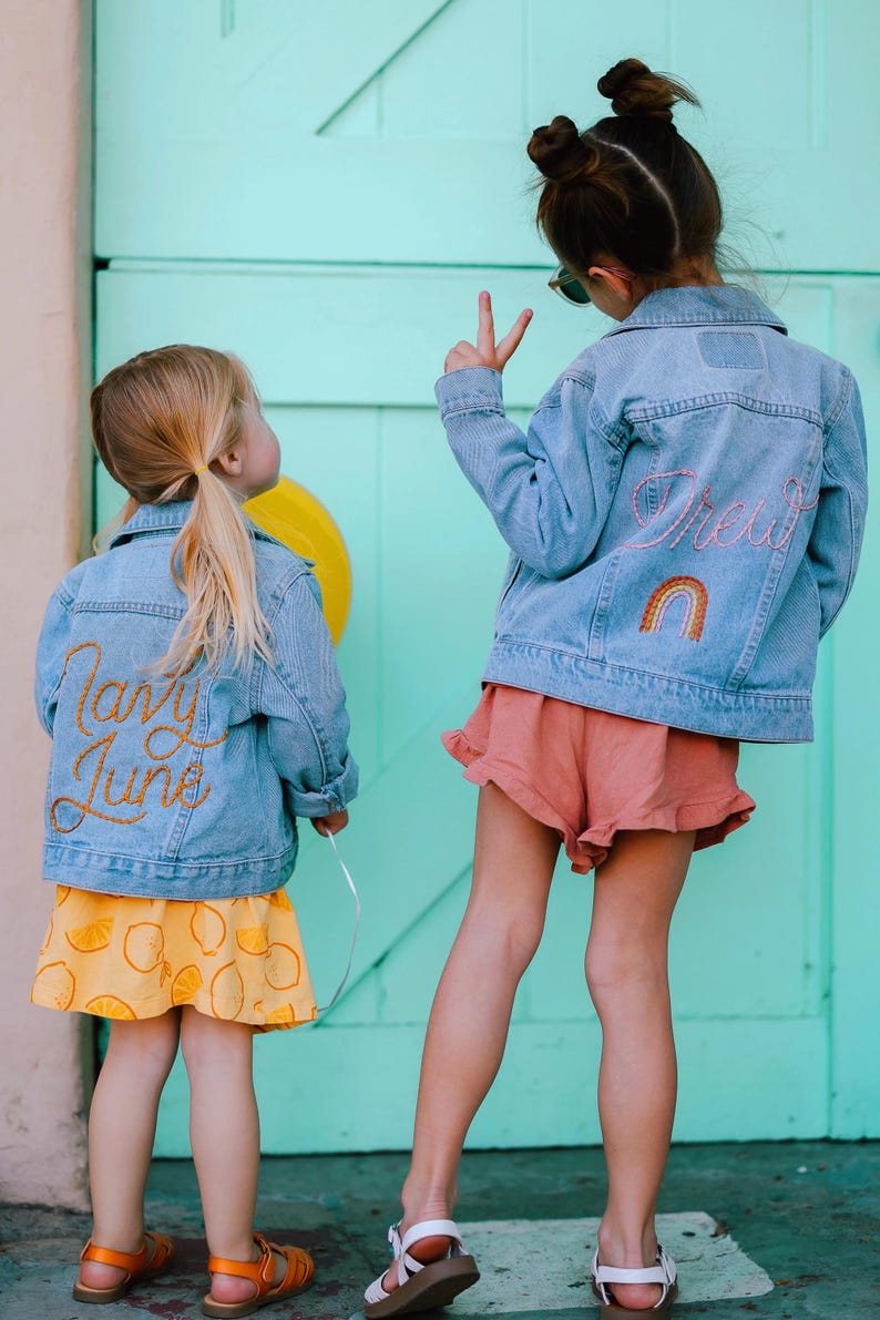 Puede incluir: Dos ni&ntilde;as peque&ntilde;as con chaquetas vaqueras con dise&ntilde;os bordados. La ni&ntilde;a de la izquierda lleva una falda amarilla con limones y el texto "Candy June" bordado en su chaqueta. La ni&ntilde;a de la derecha lleva una falda rosa y el texto "Drew" y un arco&iacute;ris bordado en su chaqueta.