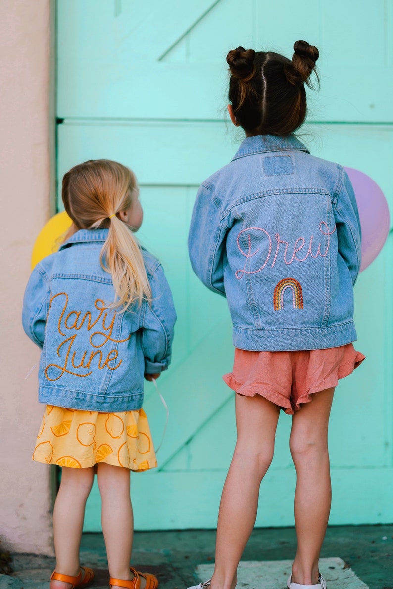 May include: Two children wearing denim jackets with embroidered names and a rainbow on the back. The girl on the left is wearing a yellow skirt with lemons on it. The girl on the right is wearing a coral colored skirt.