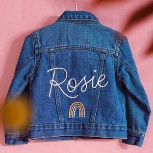 Puede incluir: Una chaqueta vaquera azul con el nombre "Rosie" bordado en la espalda con hilo blanco. Un arco&iacute;ris est&aacute; bordado debajo del nombre.