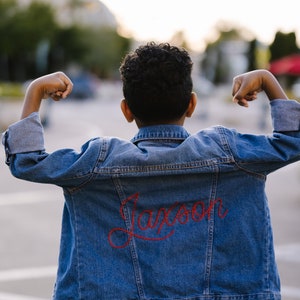 May include: A young person wearing a blue denim jacket with the name "Jackson" embroidered in red on the back. The person is standing with their arms raised in a pose of strength.