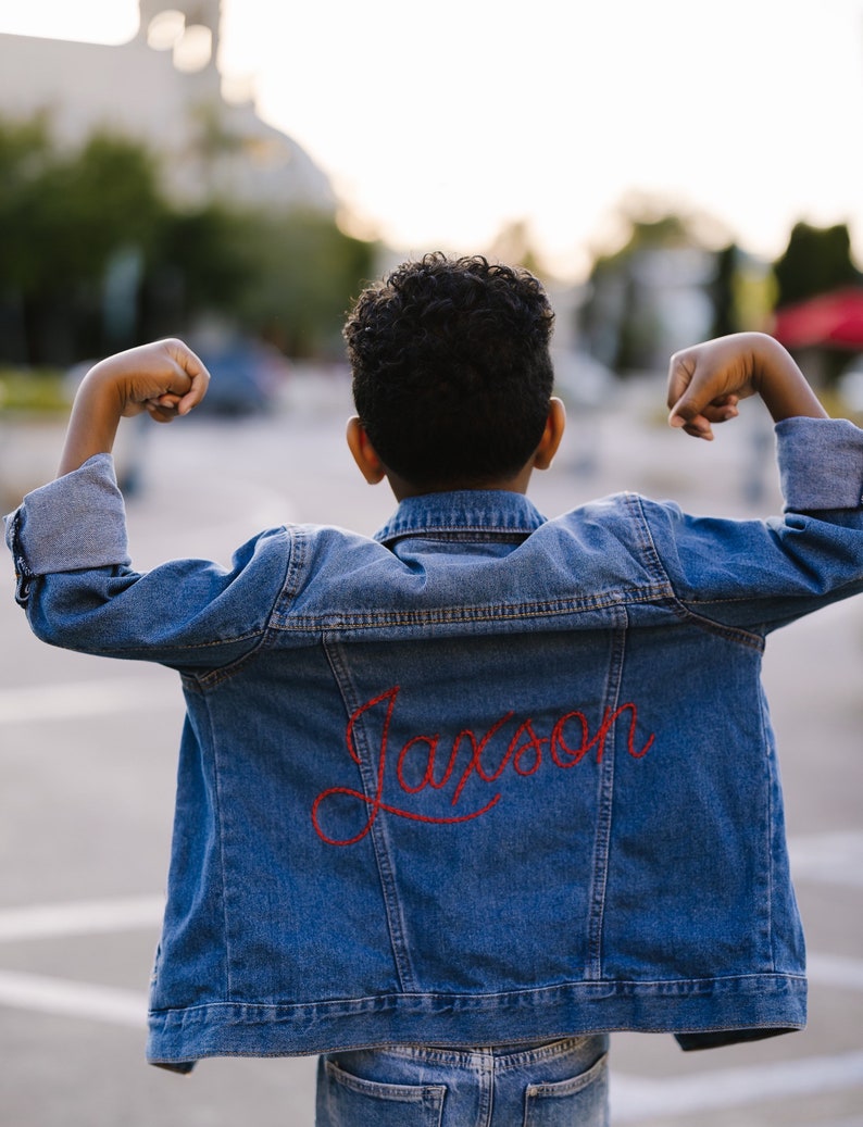 May include: A young person wearing a blue denim jacket with the name "Jackson" embroidered in red on the back. The person is standing with their arms raised in a pose of strength.